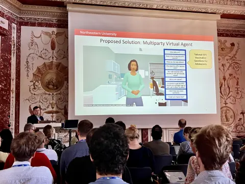 Audience in an ornate frescoed hall watching a talk about a multiparty virtual agent for HPV vaccination