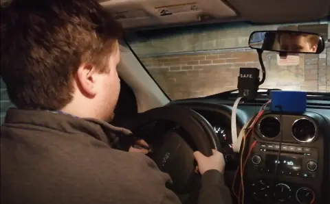 Driver at a steering wheel monitored by a dashboard-mounted S.A.F.E. camera