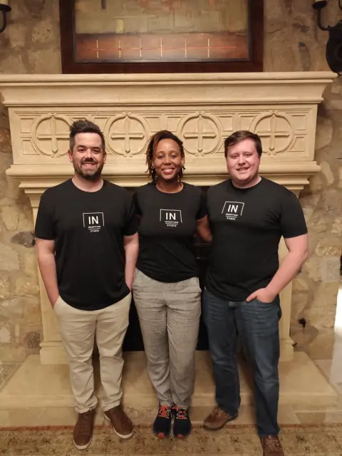 Three people in matching Inception Studio T-shirts smiling in front of a carved stone fireplace