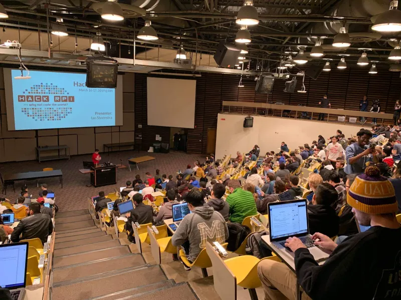 Packed auditorium of students on laptops facing a HackRPI opening slide